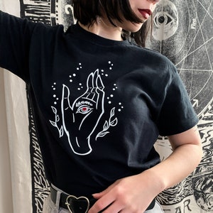 May include: Black t-shirt with a white graphic of a hand with an eye in the palm. The hand is surrounded by small white dots and stylized leaves.