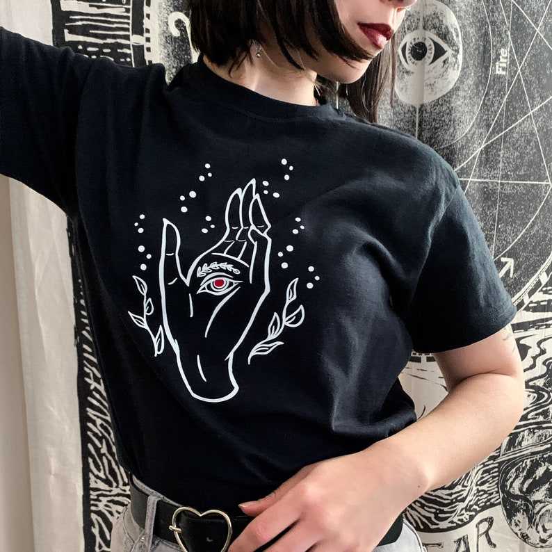 May include: Black t-shirt with a white graphic of a hand with an eye in the palm. The hand is surrounded by small white dots and stylized leaves.