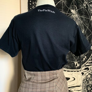 May include: Black t-shirt with the text "ThePinWitch" printed on the back. The t-shirt is being modeled on a person wearing a plaid skirt.