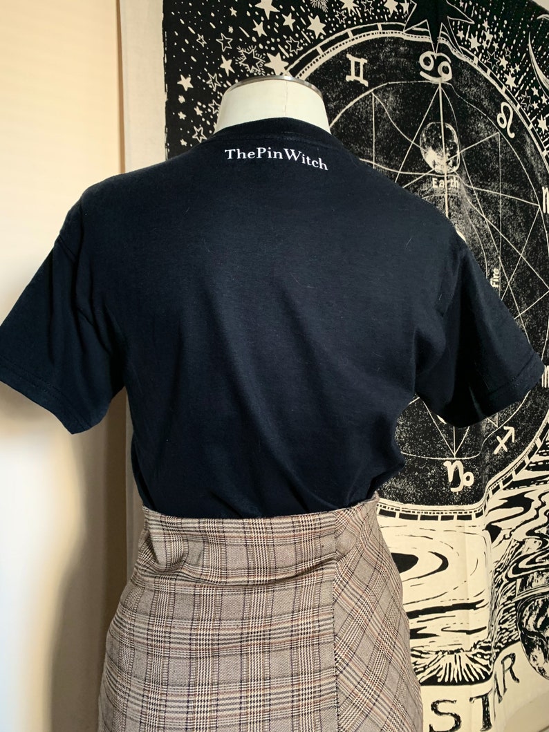 May include: Black t-shirt with the text "ThePinWitch" printed on the back. The t-shirt is being modeled on a person wearing a plaid skirt.