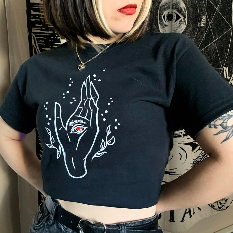 May include: Black cropped t-shirt with a white graphic of a hand with an eye in the palm. The eye is red.