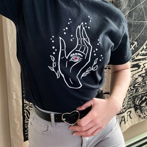 May include: A black t-shirt with a white graphic of a hand with an eye in the palm. The hand is surrounded by small white dots and leaves. The t-shirt is being worn by a person with a black belt and a heart-shaped buckle.
