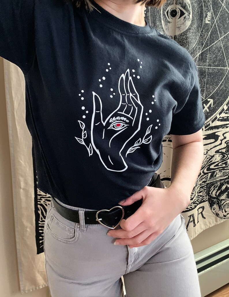 May include: A black t-shirt with a white graphic of a hand with an eye in the palm. The hand is surrounded by small white dots and leaves. The t-shirt is being worn by a person with a black belt and a heart-shaped buckle.