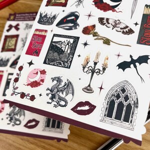 May include: A sheet of stickers featuring a variety of gothic and fantasy themed designs. The stickers include a dragon, a castle, a candle holder, a moth, a crown, a window, and a heart. The stickers are printed on white paper with a red border.