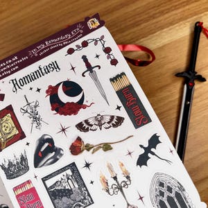 May include: A sheet of stickers with a romantic fantasy theme. The stickers feature a variety of images, including a book, a crown, a sword, a moon, a butterfly, a dragon, a candle, and a gothic window. The stickers are printed on white paper with black outlines and red accents.