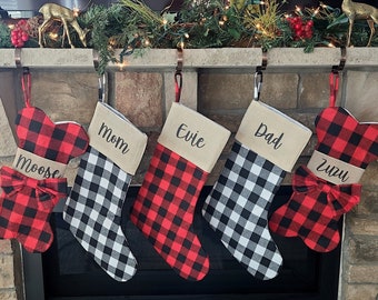 Op de afbeelding: Vijf rode en zwarte kerstsokken met buffalo plaid met gepersonaliseerde namen. De sokken hangen aan een bakstenen schoorsteenmantel met een groene krans en lampjes.