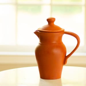 Può includere: Una brocca color terracotta con coperchio e manico. La brocca ha un corpo arrotondato e un beccuccio per versare. Il manico è curvo e il coperchio ha un piccolo pomello. La brocca è appoggiata su una superficie bianca.