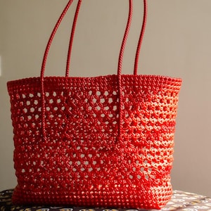 May include: A vibrant red tote bag with a woven, open-weave design. The bag features two long, matching red handles. The bag's construction allows for visibility of the interior. The bag is made of a shiny, durable material.