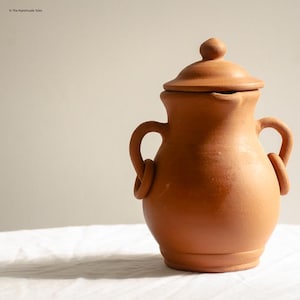 May include: A terracotta-colored ceramic jar with a lid. The jar has two loop handles and a small spout. The lid has a small knob on top. The jar is sitting on a white surface.