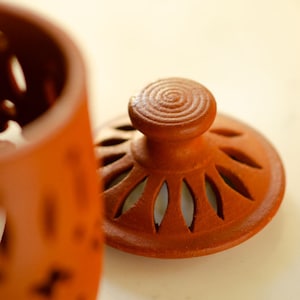 May include: A terracotta jar with a lid. The lid has a decorative design with cutouts and a knob on top. The jar is a reddish-brown colour.