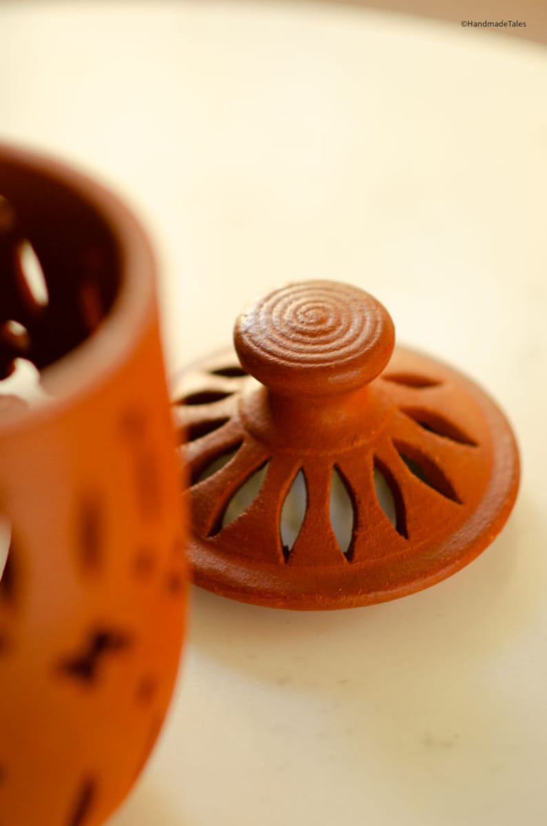 May include: A terracotta jar with a lid. The lid has a decorative design with cutouts and a knob on top. The jar is a reddish-brown colour.