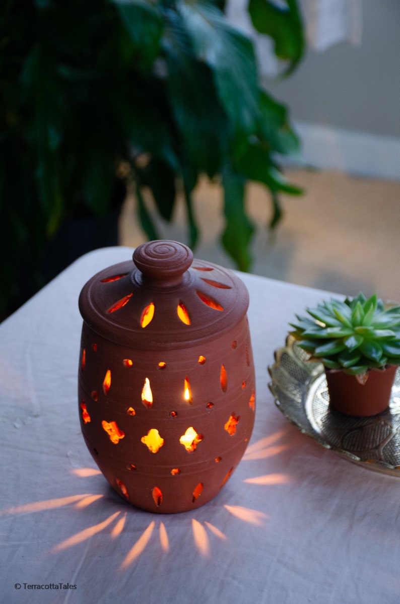 May include: A terracotta candle holder with intricate cutouts, lit with a candle inside, casting a warm glow. The holder is on a white tablecloth with a small potted succulent in the background.