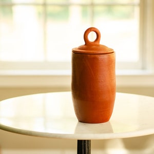 May include: A terracotta-colored ceramic jar with a lid and a loop handle. The jar is cylindrical with a slightly rounded base. The lid has a small loop for easy removal. The jar sits on a white marble table.