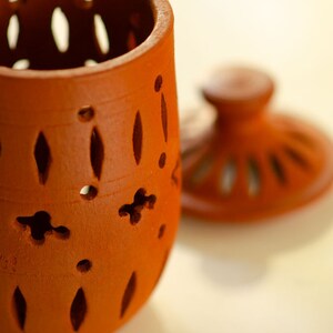 May include: A terracotta jar with a lid, both featuring intricate cut-out designs. The jar is a reddish-brown colour and has a round shape. The lid is slightly smaller than the jar and has a raised knob on top.