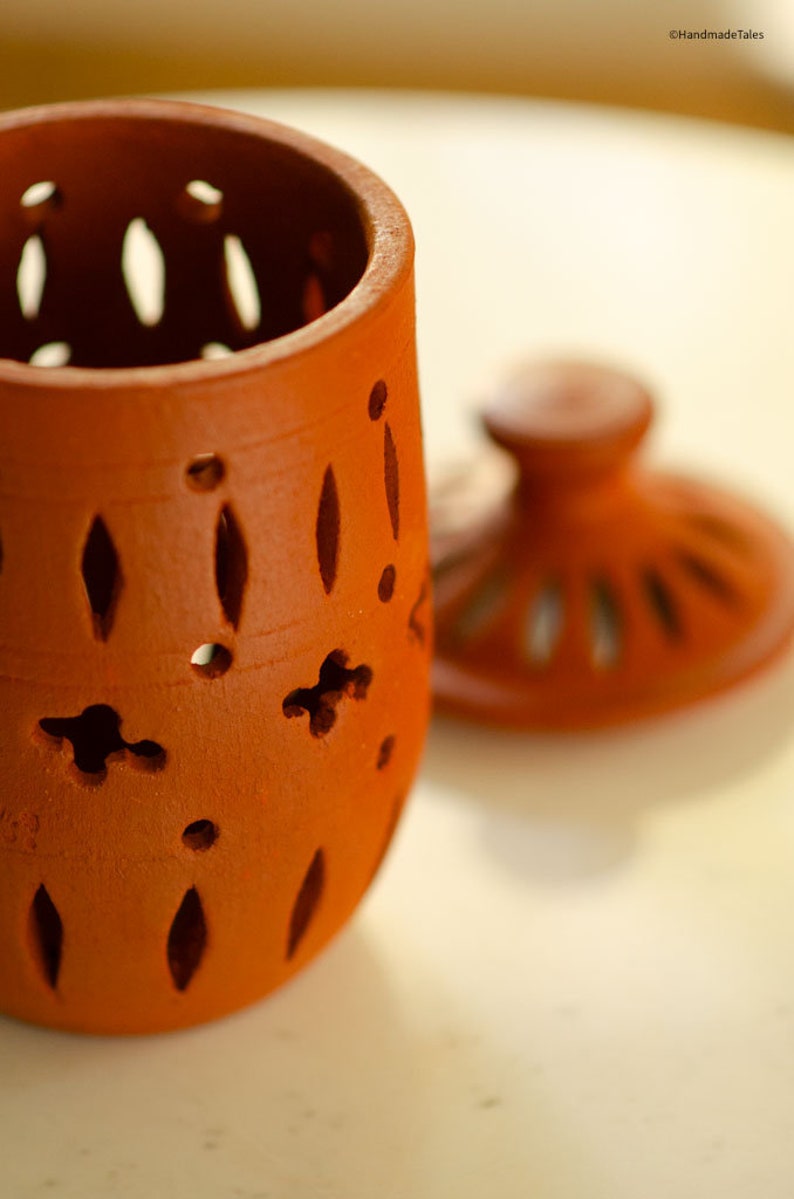 May include: A terracotta jar with a lid, both featuring intricate cut-out designs. The jar is a reddish-brown colour and has a round shape. The lid is slightly smaller than the jar and has a raised knob on top.