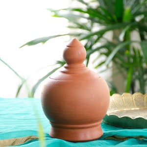 Peut inclure: Un pot artisanal de couleur terre cuite, avec un corps arrondi et un couvercle conique. Le pot est posé sur une surface turquoise, avec un bord doré visible. Un plat décoratif en laiton est partiellement visible.