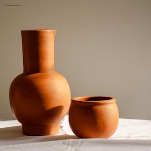 Puede incluir: Dos jarrones de terracota. Uno es un jarrón alto y redondeado con un cuello estrecho. El otro es una maceta pequeña y redonda. Ambos son de un color marrón anaranjado cálido y terroso. Los jarrones están sobre una superficie blanca.