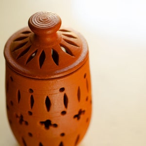 May include: A terracotta jar with a lid, featuring a decorative design of cut-out circles and diamond shapes. The jar has a rustic, handmade look.