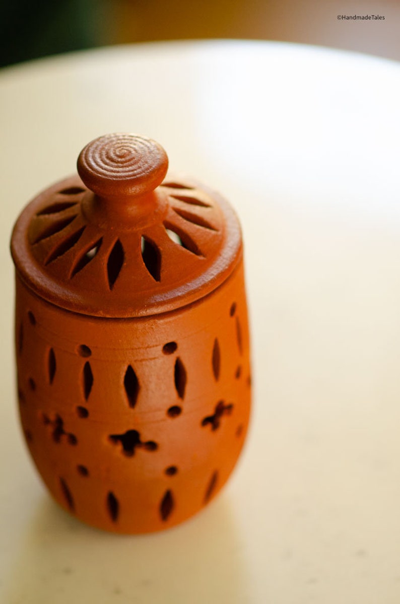 May include: A terracotta jar with a lid, featuring a decorative design of cut-out circles and diamond shapes. The jar has a rustic, handmade look.