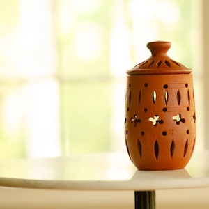 May include: A terracotta jar with a lid, featuring intricate cut-out designs. The jar is sitting on a white table top.
