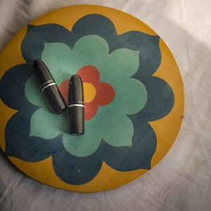 Puede incluir: Dos tubos de pintalabios cilíndricos negros con el logotipo de MAC impreso en plata, colocados sobre un azulejo decorativo circular con un patrón floral estilizado en tonos amarillo mostaza, azul cerceta, azul oscuro y una flor central roja y amarilla.