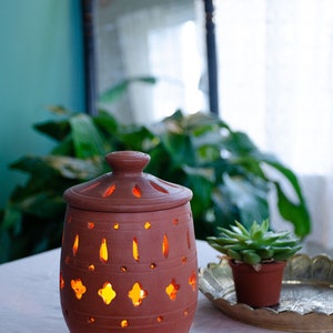 May include: A terracotta candle holder with intricate cutouts, lit from within, casting a warm glow. The holder is on a white table with a small potted succulent plant and a decorative tray.