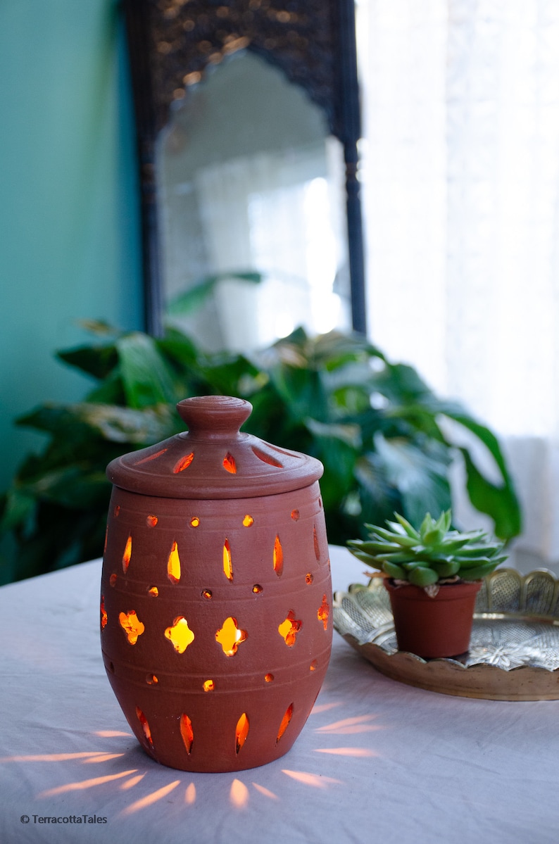 May include: A terracotta candle holder with intricate cutouts, lit from within, casting a warm glow. The holder is on a white table with a small potted succulent plant and a decorative tray.