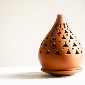 Op de afbeelding: Een terracotta-kleurige, kegelvormige kandelaar met driehoekige en cirkelvormige uitsparingen. De basis heeft een bijpassende terracotta-kleur en is rond. The Handmade Tales staat linksboven afgedrukt.