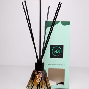 May include: A brown glass diffuser bottle with black reeds and a gold leaf charm. The bottle has a faceted design and is sitting on a white surface. The bottle is in front of a green and white box with the text "AE" and "www.affirmationeffect.com".