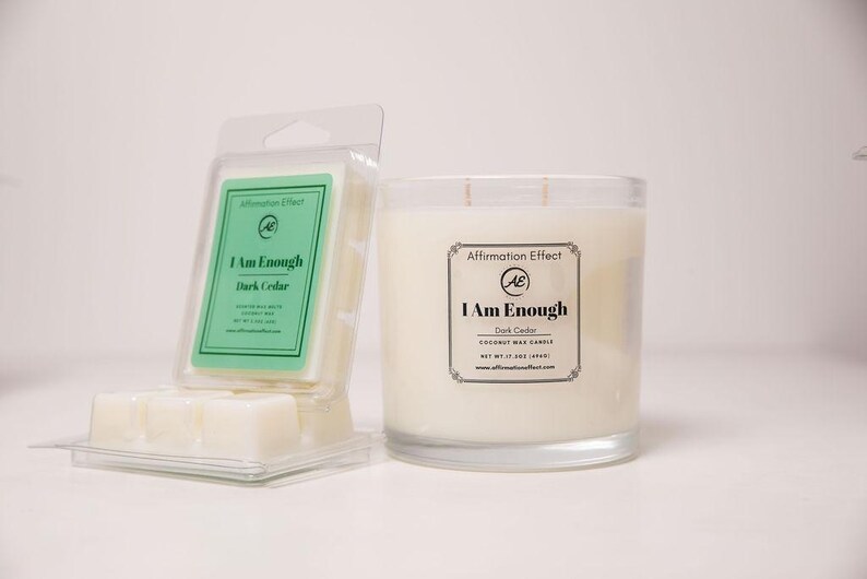 May include: A candle and wax melts set. The candle is in a clear glass jar with a white wax and two wicks. The wax melts are in a clear plastic container. Both products have labels that read "I Am Enough Dark Cedar".