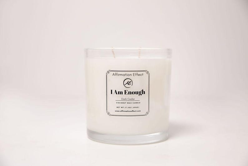 May include: A clear glass candle jar filled with white wax. The label reads "I Am Enough" and "Dark Cedar Coconut Wax Candle". The candle weighs 17.5 oz (496g). The brand name is "Affirmation Effect".
