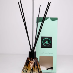 Pode incluir: Uma garrafa de difusor de vidro marrom com juncos pretos e um amuleto de folha de ouro. A garrafa tem um design facetado e está sobre uma superfície branca. A garrafa está na frente de uma caixa verde e branca com o texto "AE" e "www.affirmationeffect.com".