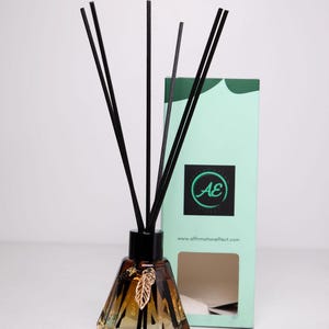 Puede incluir: Una botella de difusor de vidrio marrón con cañas negras y un dije de hoja de oro. La botella tiene un diseño facetado y está sobre una superficie blanca. La botella está delante de una caja verde y blanca con el texto "AE" y "www.affirmationeffect.com".