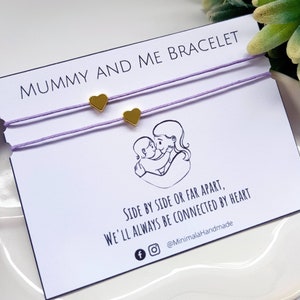 May include: Two matching purple string bracelets with gold heart charms. The bracelets are displayed on a white card with the text "Mummy and Me Bracelet" and "Side by side or far apart, we'll always be connected by heart" and the social media handle "@MinimalaHandmade".