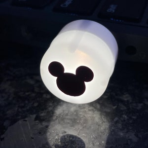 May include: White round candle with a black Mickey Mouse silhouette on the front. The candle is lit and casting a glow.