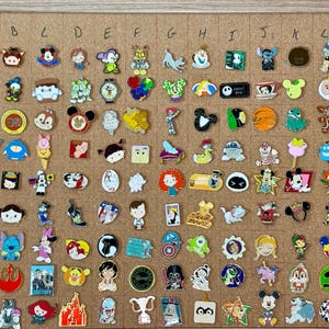 May include: A collection of various colorful enamel pins arranged in a grid on a cork board. The pins feature diverse cartoon characters, symbols, and shapes, organized by rows numbered one through nine and columns labeled A through M.