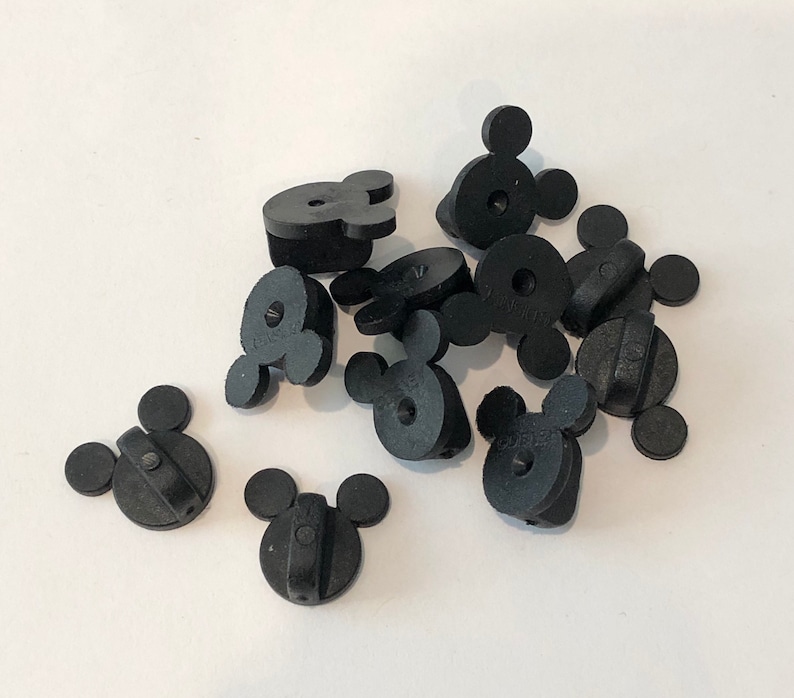 May include: A group of ten black plastic Mickey Mouse head shaped pin backs. The pin backs are all the same size and shape, and they are arranged in a random pattern.