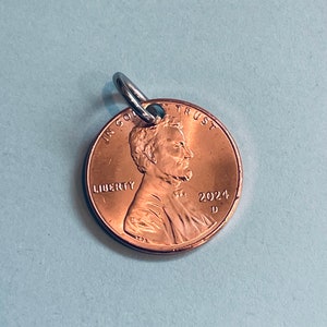 May include: A copper-colored US penny with a silver ring attached to the top. The coin features a profile of Abraham Lincoln on the front, with the words "IN GOD WE TRUST", "LIBERTY", and "2024 D".