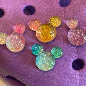 May include: Five colorful glitter Mickey Mouse head charms in various colors, including pink, yellow, green, and blue. The charms are on a purple background.
