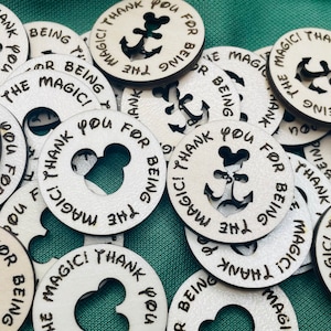 May include: A collection of round wooden tokens with white backgrounds and black lettering. The tokens feature different designs, including anchors, Mickey Mouse ears, and the text "Thank you for being the magic!"