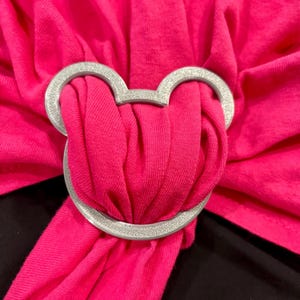 May include: A silver-colored Mickey Mouse-shaped accessory adorns a vibrant pink fabric. The accessory is circular with a textured surface, and the pink fabric is gathered and draped, creating a layered effect. The background is black.