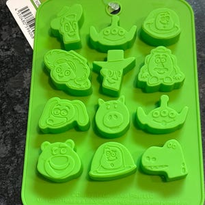 May include: A green silicone chocolate mold featuring various Toy Story character designs. The mold includes shapes of Woody, Buzz Lightyear, and other characters. The mold is rectangular and has a hole for hanging.
