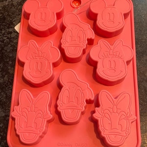May include: Pink silicone mold with eight cavities shaped like Mickey Mouse, Minnie Mouse, and Donald Duck. The mold is labeled "Disney DAISO".