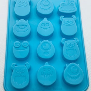 May include: A light blue silicone chocolate mold featuring various cartoon monster designs. The mold has 12 cavities, each with a different monster face. The mold is rectangular and includes the text "©Disney/Pixar DAISO".