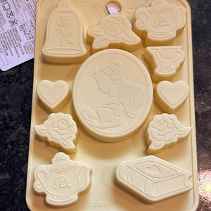 May include: A light yellow silicone chocolate mold featuring various shapes, including a rose in a bell jar, a rose, a teapot, a teacup, hearts, and a book. The mold also includes a circular design with a silhouette of a woman. The Disney logo is visible.