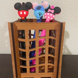 May include: A wooden pencil holder with a grid pattern. Three pens with cartoon character toppers are in the holder. The toppers are Mickey Mouse, Minnie Mouse, and a blue character with a paw print.