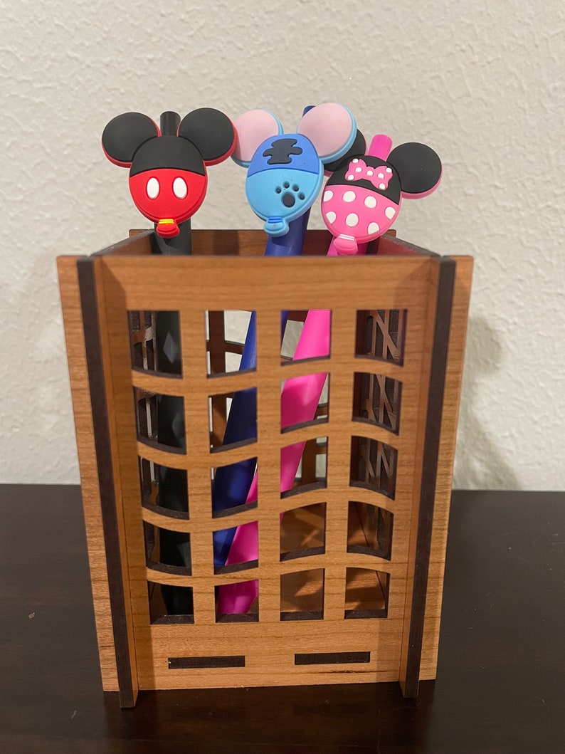 May include: A wooden pencil holder with a grid pattern. Three pens with cartoon character toppers are in the holder. The toppers are Mickey Mouse, Minnie Mouse, and a blue character with a paw print.