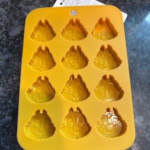 May include: A yellow silicone mold featuring Chip and Dale faces. The mold has 12 cavities, each shaped like the cartoon chipmunks. The mold is designed for making chocolates, candies, or ice cubes. A white tag is attached to the mold.