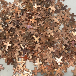 May include: A collection of small, copper-colored metal crosses scattered across a surface. The crosses have a simple, four-arm design and a slightly textured appearance. The image is a close-up, showing the details of the individual crosses.