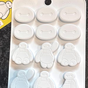 May include: White silicone mold featuring Baymax from Big Hero 6. The mold has 12 cavities, including oval shapes and Baymax character designs. A yellow tag with Japanese text is attached to the mold.